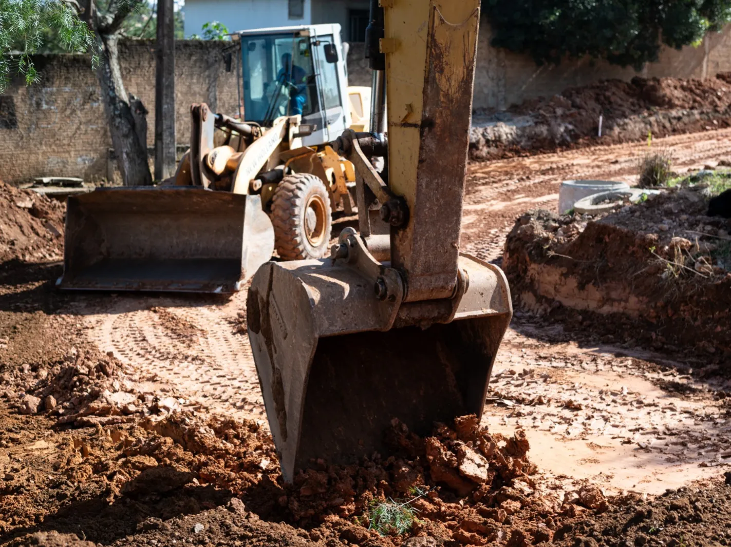 Homem em cadeira de rodas em terreno de terra com máquina amarela de construção ao fundo.